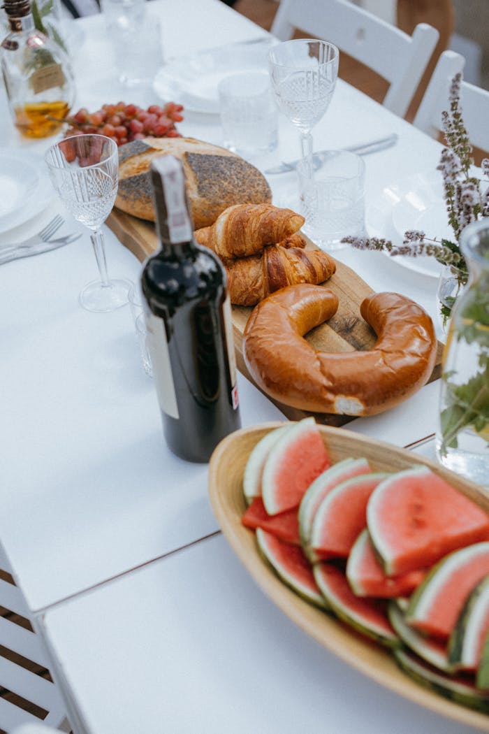 gallery-01 An elegant outdoor dining table set with wine, bread, croissants, and watermelon slices, perfect for a celebration.